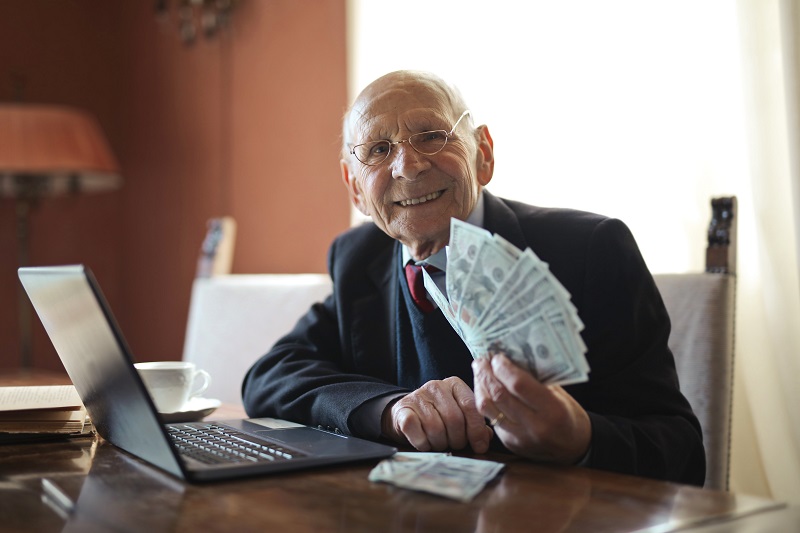 An old man holding cash and grinning, while sitting in front of a computer