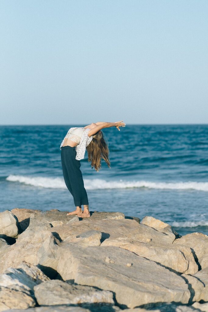 A young caucasian woman doing yoga on top of rocks with a beach in the background.