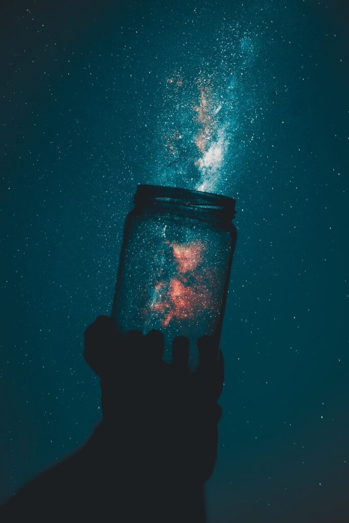 Living In A Simulation - An image showing a beautiful night time shot of a galaxy with a person holding a jar in front of it. The refraction creates the illusion that the galaxy flows out of the glass jar.