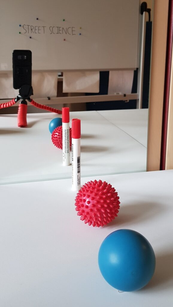 In this photo, a blue ball, a red ball, and a red marker are placed in series in front of a mirror. The blue ball is closest to you, the red ball is in the middle, and the red marker is the farthest from you. But in the reflection, the red marker appears closest to you, the red ball appears in the middle, and the blue ball appears the farthest from you.