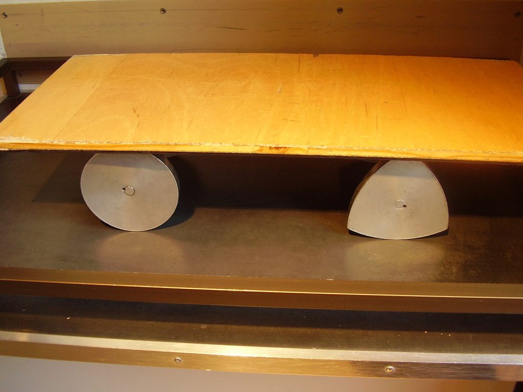 How To Really Benefit From Curves Of Constant Width? - A wooden plank placed on top of two differently shaped objects (Image from WikiCC). One object features a circular corss-section whereas the other object features a curved triangular cross-section.
