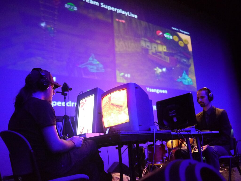 How To Use Video Games To Solve Scientific Problems — An image showing two speedrunners playing The Legend of Zelda (2013). The room looks dark and is lit in purple with computers and broadcast systems displayed.